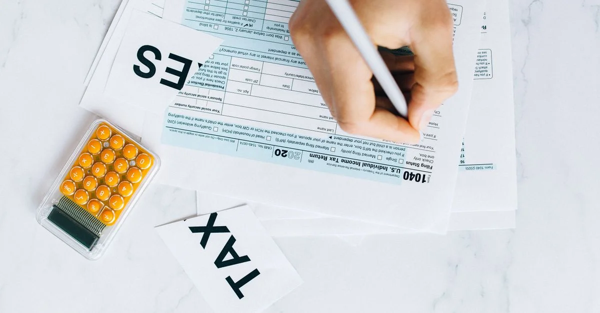 Gros plan sur une main écrivant sur un formulaire fiscal avec une calculatrice à proximité sur une surface blanche.