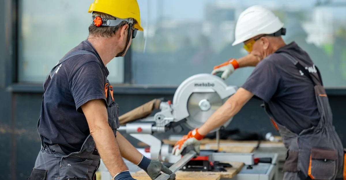 Deux ouvriers du bâtiment coupant une poutre métallique avec une scie circulaire, portant des équipements de sécurité sur le chantier.