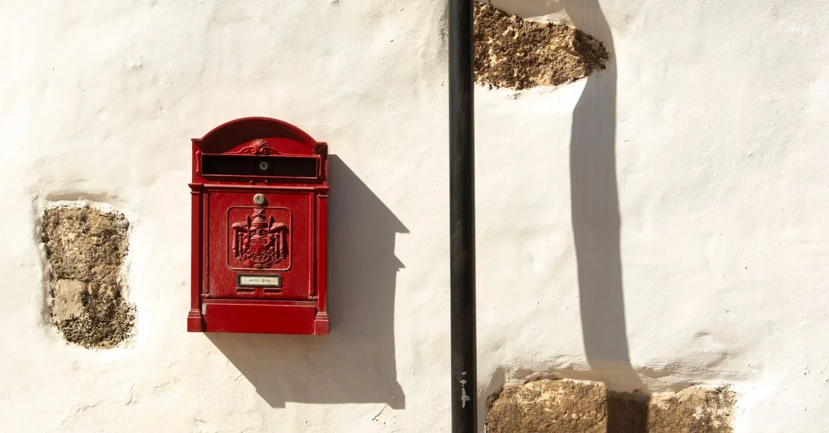 Une boîte aux lettres rouge classique sur un mur blanc texturé, mise en valeur par la lumière du soleil et les ombres.