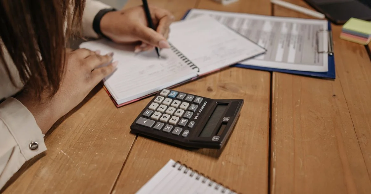 Femme prenant des notes avec une calculatrice sur un bureau en bois, concentrée sur son travail et sa comptabilité.