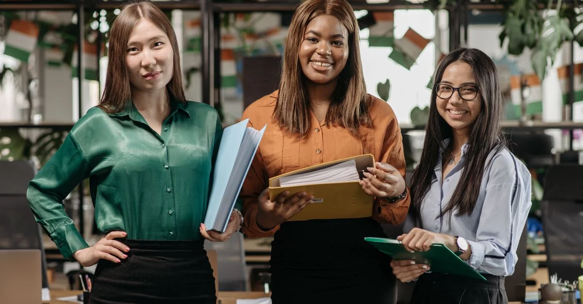Trois femmes d'affaires diverses se tenant ensemble dans un bureau, tenant des dossiers et souriant.