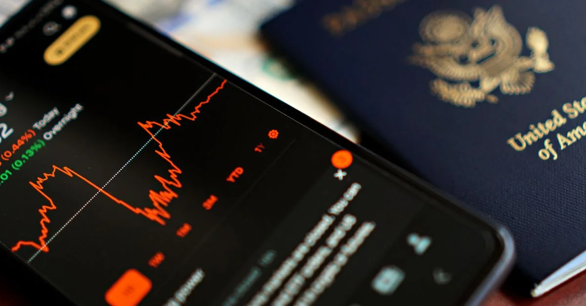 Gros plan d'un smartphone affichant des données boursières à côté d'un passeport américain, suggérant des thèmes de voyage et de finance.