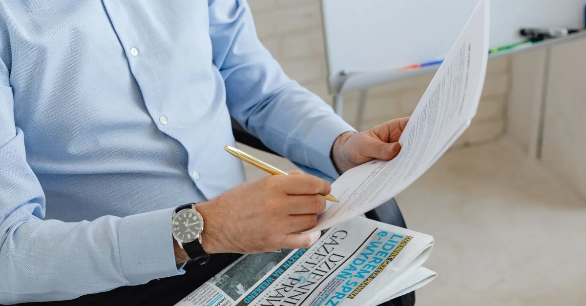 Homme d'affaires en chemise bleue examinant des documents avec un stylo sur un journal dans un bureau.