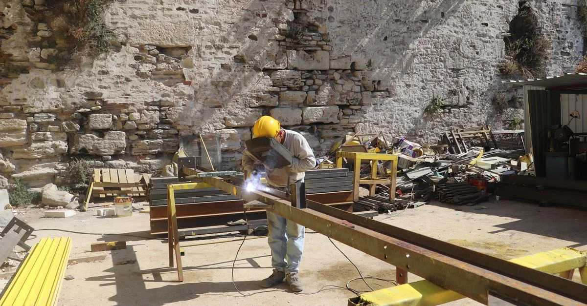 Ouvrier en équipement de protection soudant une poutre en acier sur un chantier de construction extérieur contre un mur en pierre.