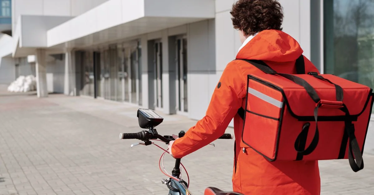 Un coursier à vélo en veste orange avec un sac de livraison sur une rue de la ville, prêt à livrer.